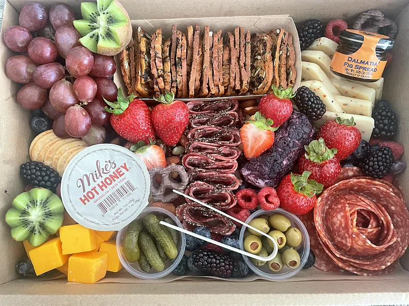 Large charcuterie box with pepper salami, a salami rose, blueberry goat cheese, hot honey, kiwi flowers, berries, olives and pickles