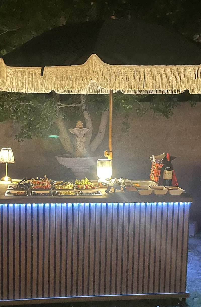 Three Hearts Grazing charcuterie cart at an evening event with a fringed umbrella, lamps and glowing under-lighting